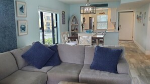 A living room with a beige sectional sofa, a dining area with a white table and chairs, and a hallway leading to a door. - Escape to your HAPPY place, luxury corner condo right on the beach! (Jensen Beach)