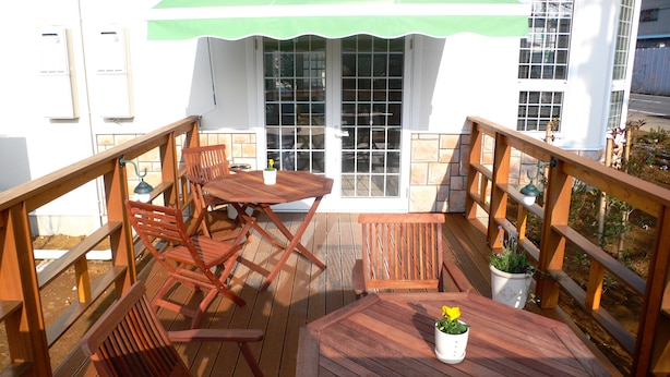 Um deck de madeira com uma mesa, cadeiras, um vaso de planta e vista para um edifício com toldo verde.