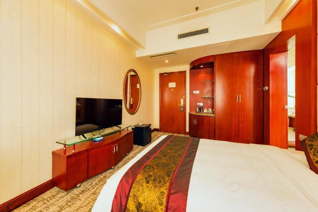A hotel room with a large bed, a flat-screen TV, a mirror, wooden wardrobe, and a small desk with a glass top.