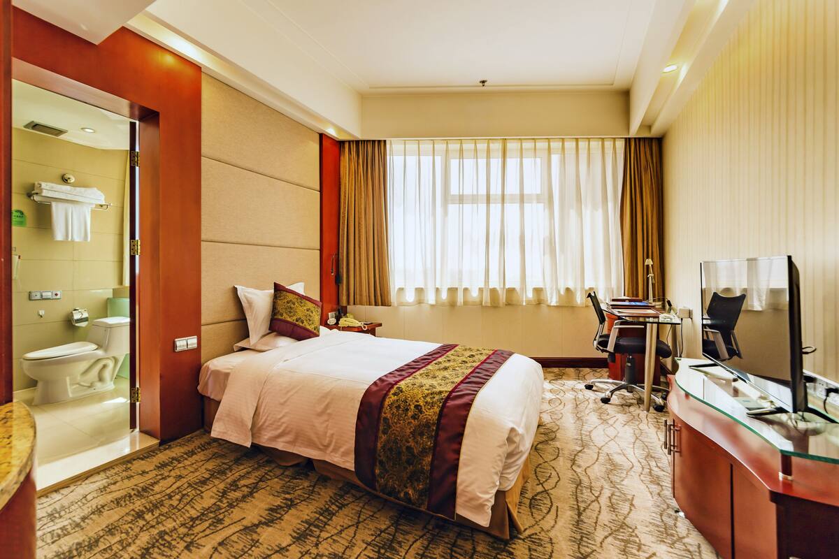 A hotel room with a bed, a desk with a computer, a chair, a window with curtains, and a bathroom with a toilet and sink.