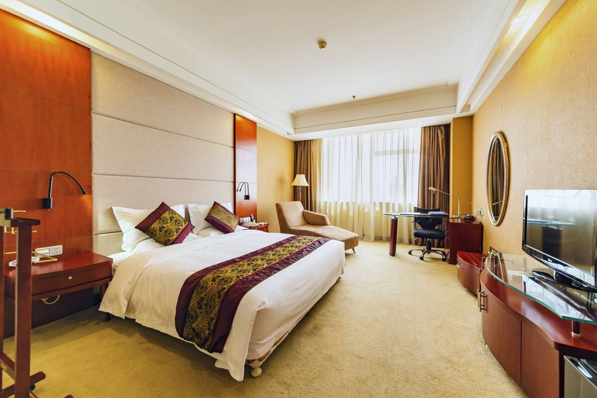 A hotel room with a large bed, a desk with a TV, a chair, and a desk lamp.