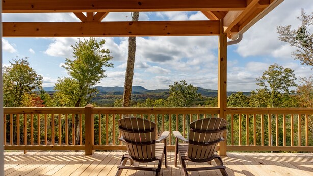 Una terraza de madera con dos sillas Adirondack y una mesa, con vistas a un paisaje montañoso.