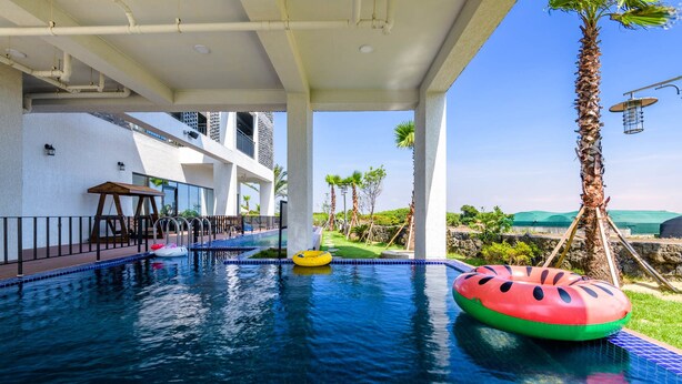 An indoor swimming pool with clear blue water, surrounded by a white wall and a covered area with a wooden table and chairs. - Modeul Spa Pension (Seogwipo)