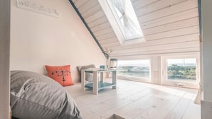 Um quarto com pé-direito alto, um domogem, uma cama com almofadas, uma mesa pequena com um vaso e janelas amplas com vista para a paisagem.