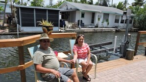 Un couple est assis sur un quai, avec une étendue d’eau et une maison en arrière-plan.