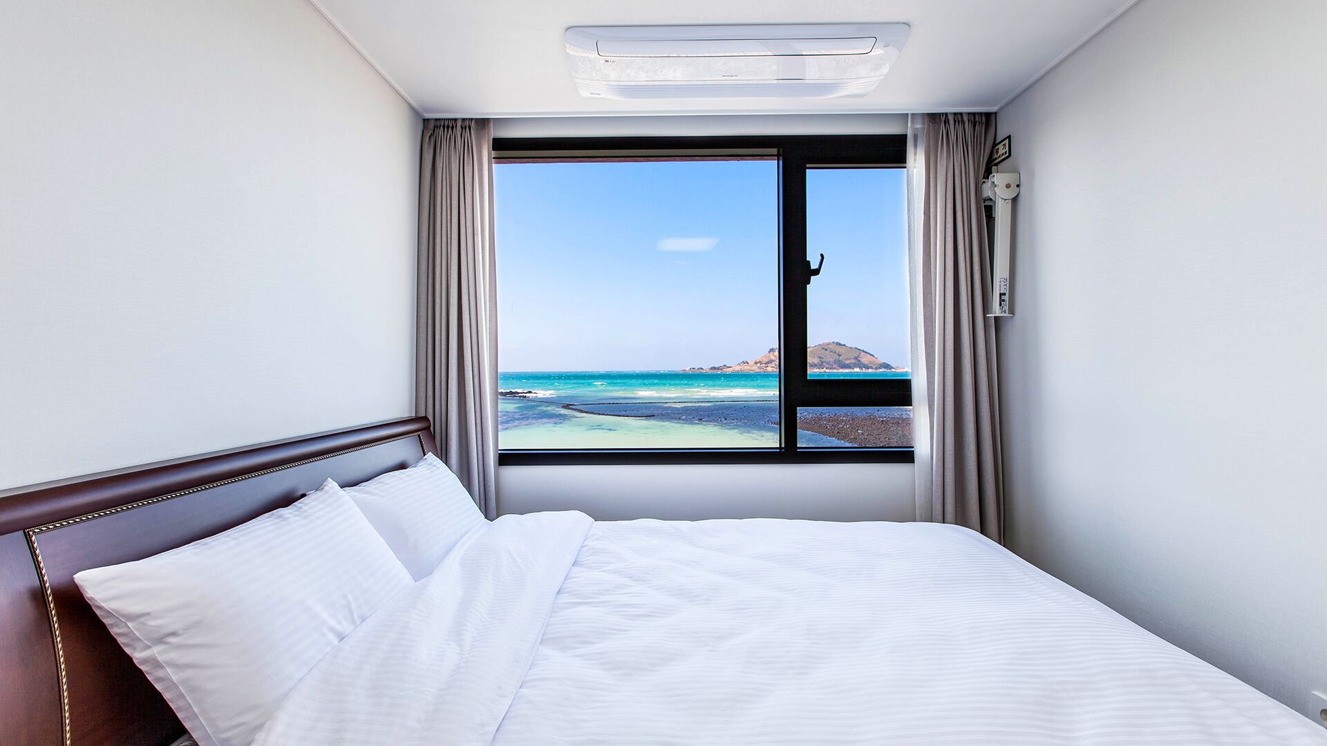 A hotel room with a bed, a window with a view of the sea, and a ceiling air conditioner.