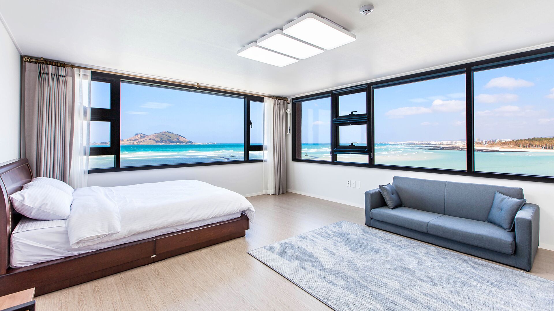A modern bedroom with a bed, a gray sofa, and large windows offering ocean views.