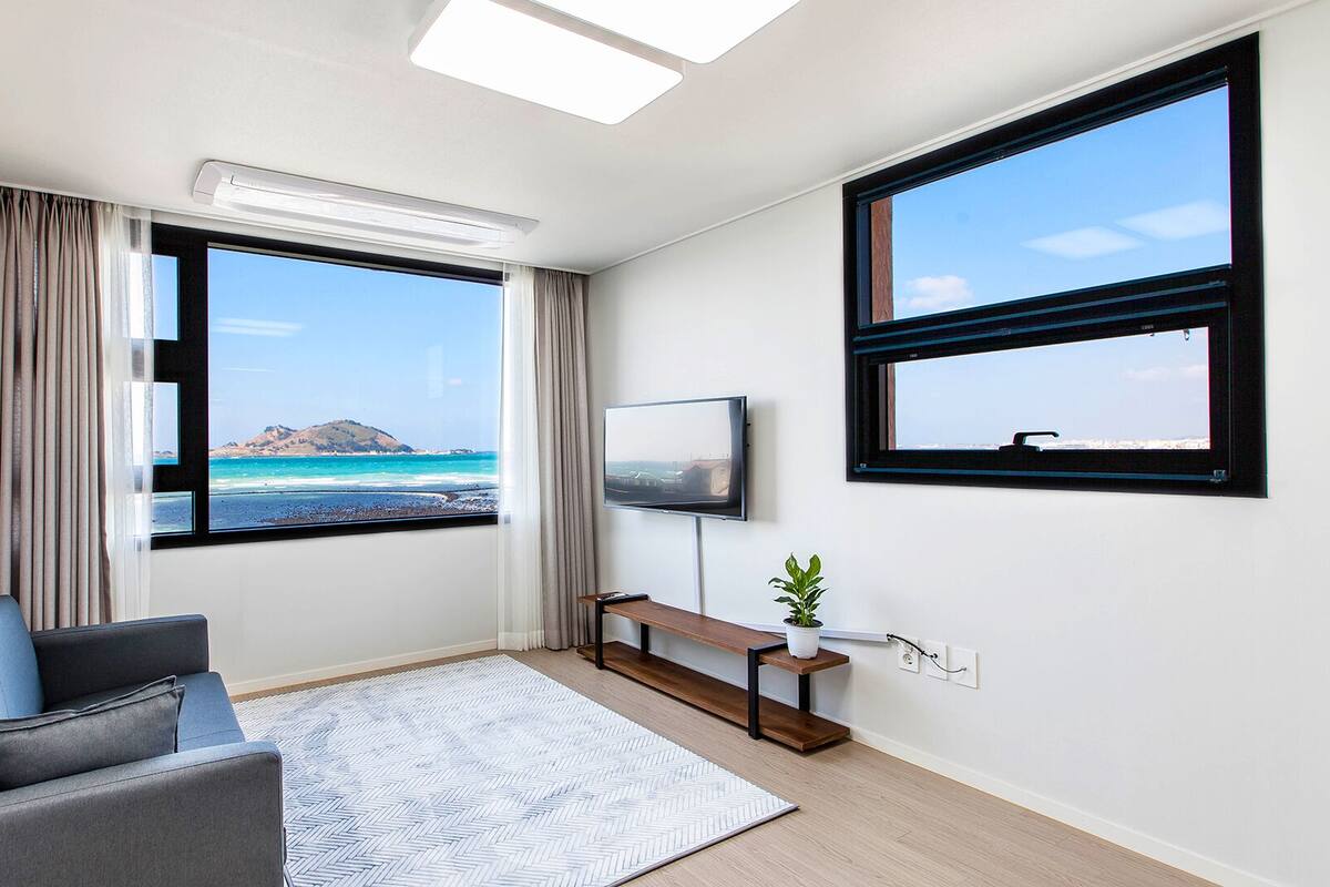 Ein modernes Zimmer mit einem großen Fenster, das Meer und Berge blickt, ein graues Sofa, ein kleiner Holztisch mit einer Pflanze und ein an der Wand befestigtes Fernsehgerät.