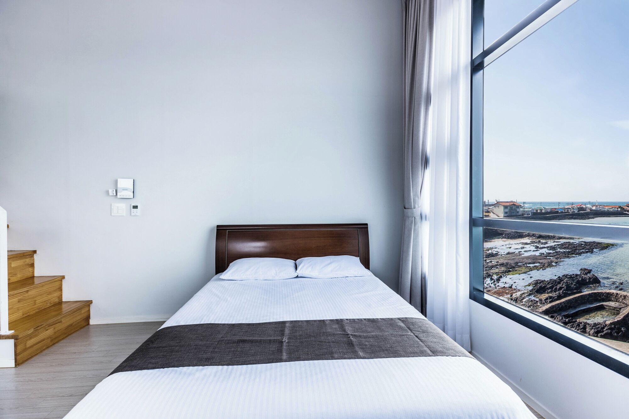 A bedroom with a large window, a bed, a wooden staircase, and a view of a coastal area.