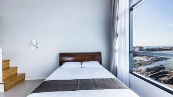 A bedroom with a large window, a bed, a wooden staircase, and a view of a coastal area.