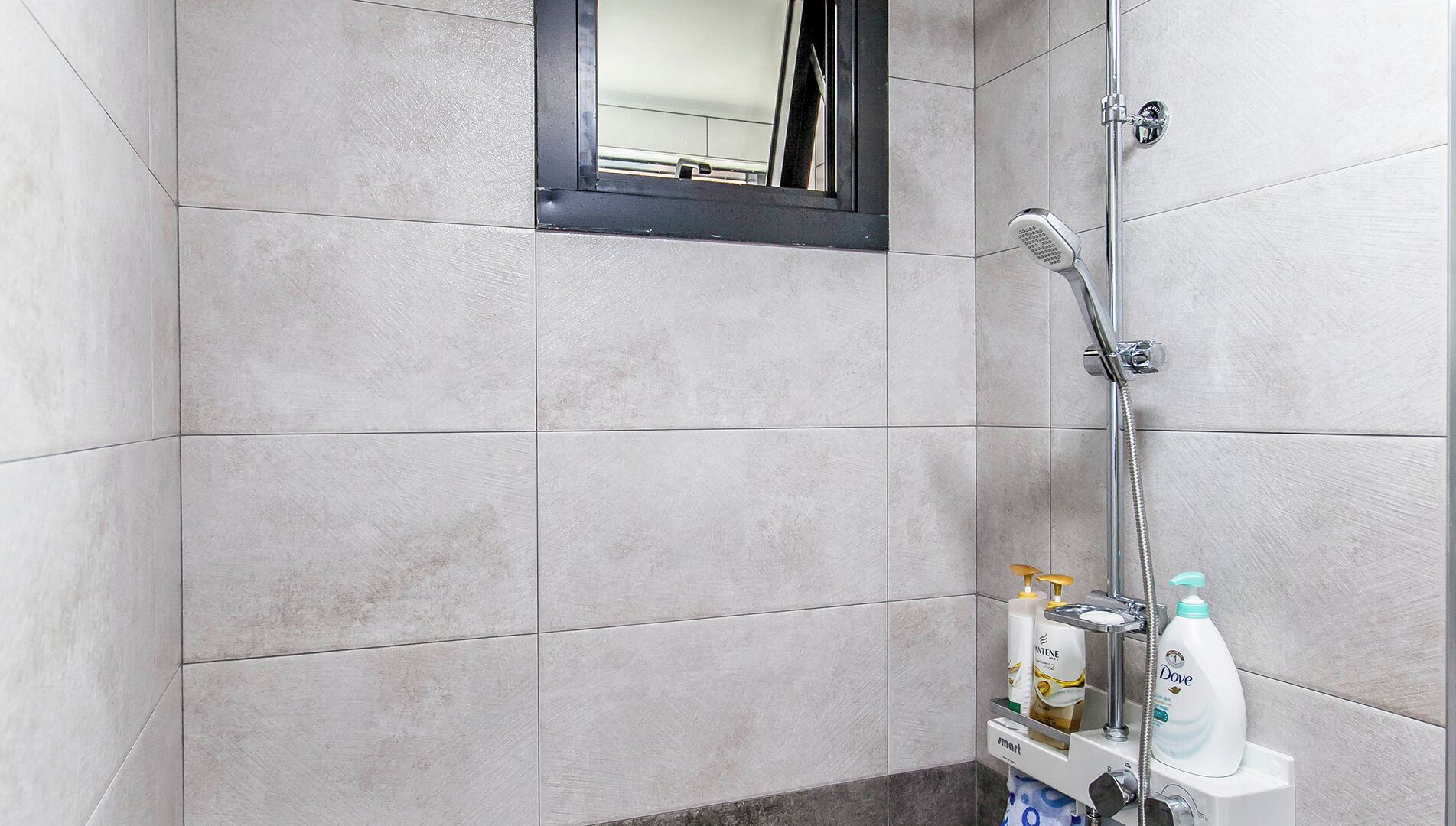 A shower area with grey tiles, a handheld showerhead, and a wall-mounted faucet. There are toiletries on a shelf.