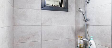 A shower area with grey tiles, a handheld showerhead, and a wall-mounted faucet. There are toiletries on a shelf.
