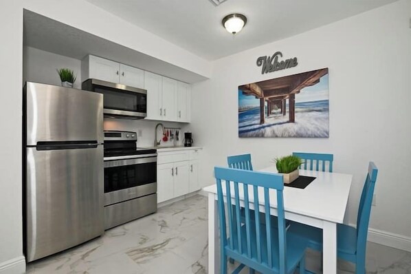 A modern kitchen with stainless steel appliances, white cabinets, a dining table with blue chairs, and a 'Welcome' sign on the wall.