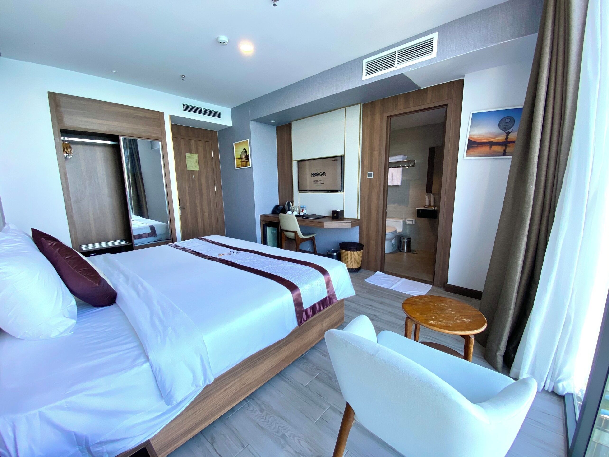 A hotel room with a bed, a desk, a chair, a small table, a mirror, and a bathroom visible through a glass door.