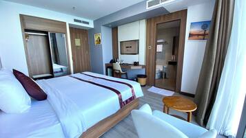 A hotel room with a bed, a desk, a chair, a small table, a mirror, and a bathroom visible through a glass door.
