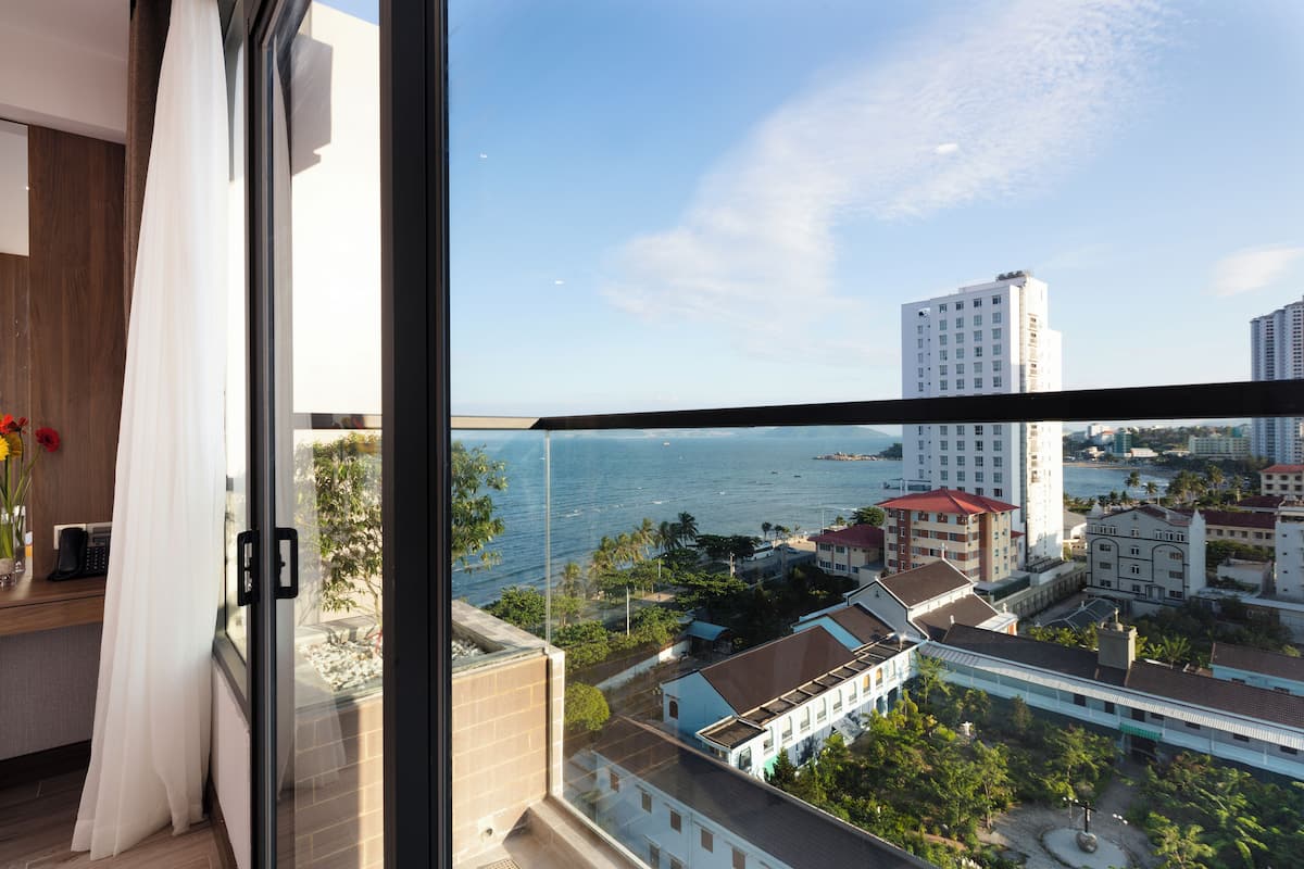 A high-rise hotel room with a large window overlooking a coastal cityscape, including a view of the sea and other buildings.