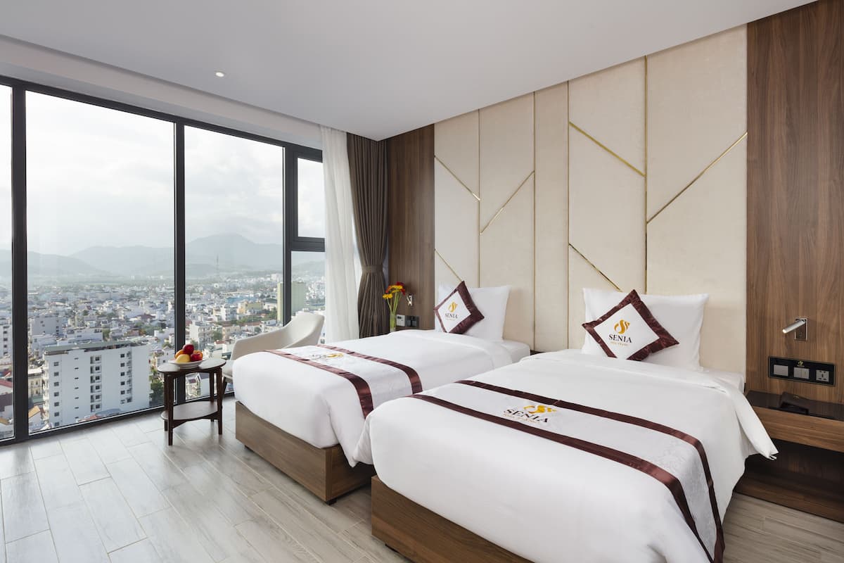 A hotel room with two beds, a large window with a city view, a small table with fruit, and a chair.