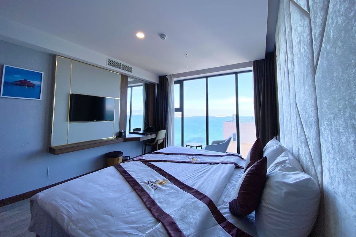 A hotel room with a large bed, a desk with a chair, a television, and a view of the sea.