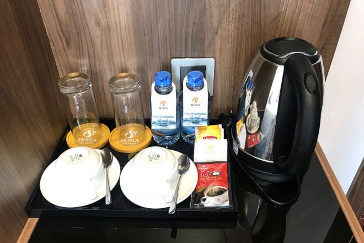 A hotel minibar with a coffee maker, two bottles of Senia water, two glasses, a spoon, a plate, and a cup of coffee.