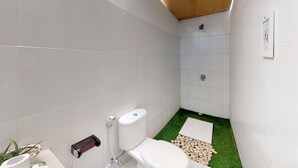 Une petite salle de bain moderne, avec des carreaux blancs, des toilettes, une douche et un petit coin de gazon artificiel.