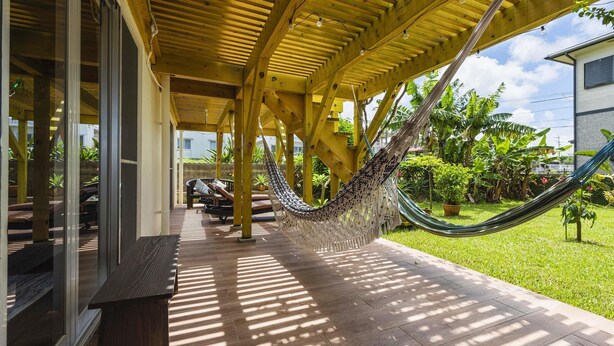 A covered patio area with a hammock, wooden flooring, and a view of a residential building. - Panorama (Yomitan)