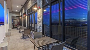 Un espace extérieur aménagé avec des tables et des chaises, de grandes fenêtres et une vue sur le paysage urbain au crépuscule.
