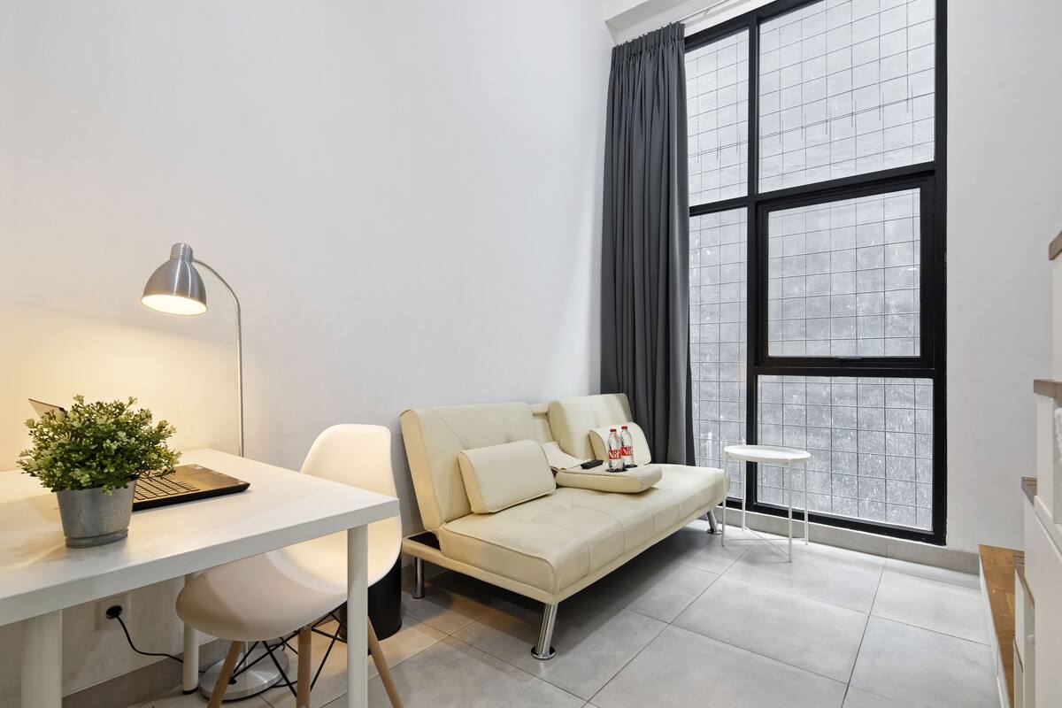 A modern room with a white desk, a beige sofa, a small side table, and a large window with curtains.