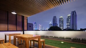 A modern outdoor seating area with wooden tables and benches, overlooking a city skyline at dusk. - Sakura Terrace Setiabudi (Jakarta)