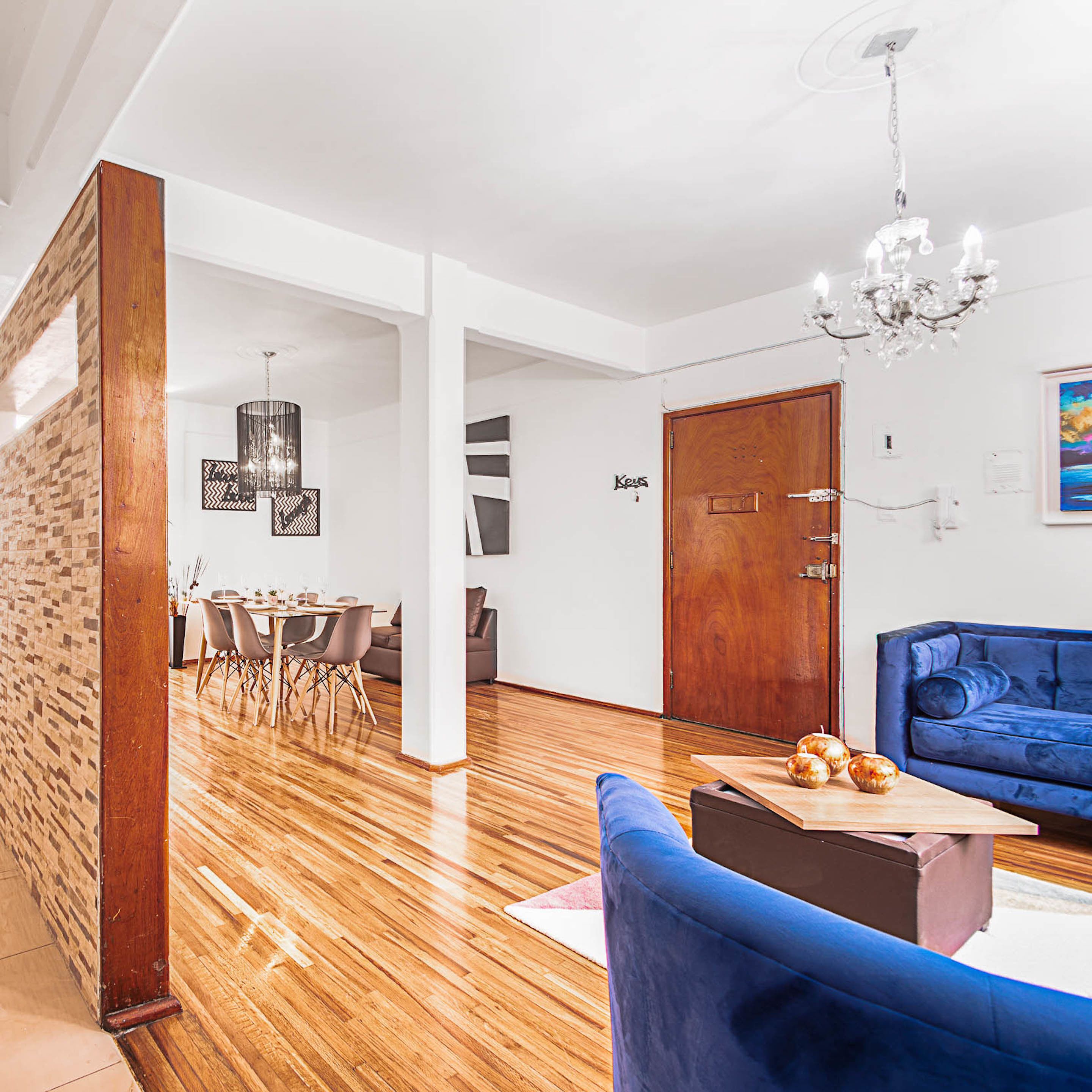 A modern living room with a wooden floor, a blue sofa, a dining area with a wooden table and chairs, and a chandelier.