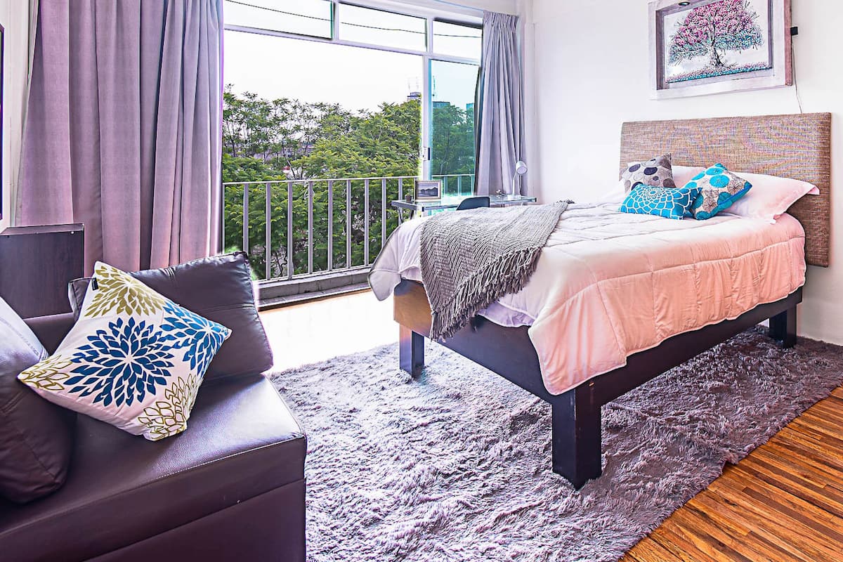 A bedroom with a bed, a chair, a balcony with curtains, and a painting on the wall.