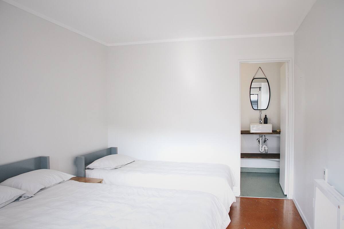 A bedroom with two beds, a bathroom with a mirror and sink, and a hallway leading to another room.