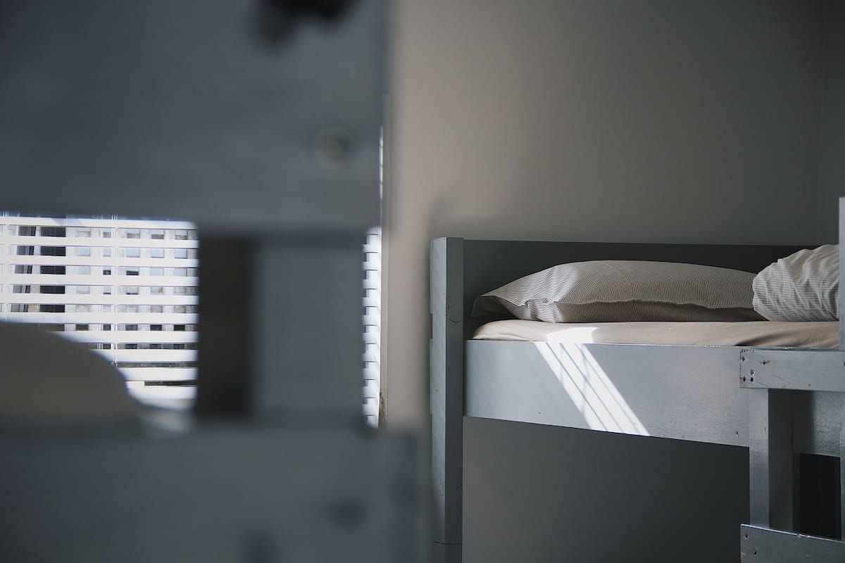 A bunk bed with a mattress and pillows, a window with blinds, and a partial view of a building outside.