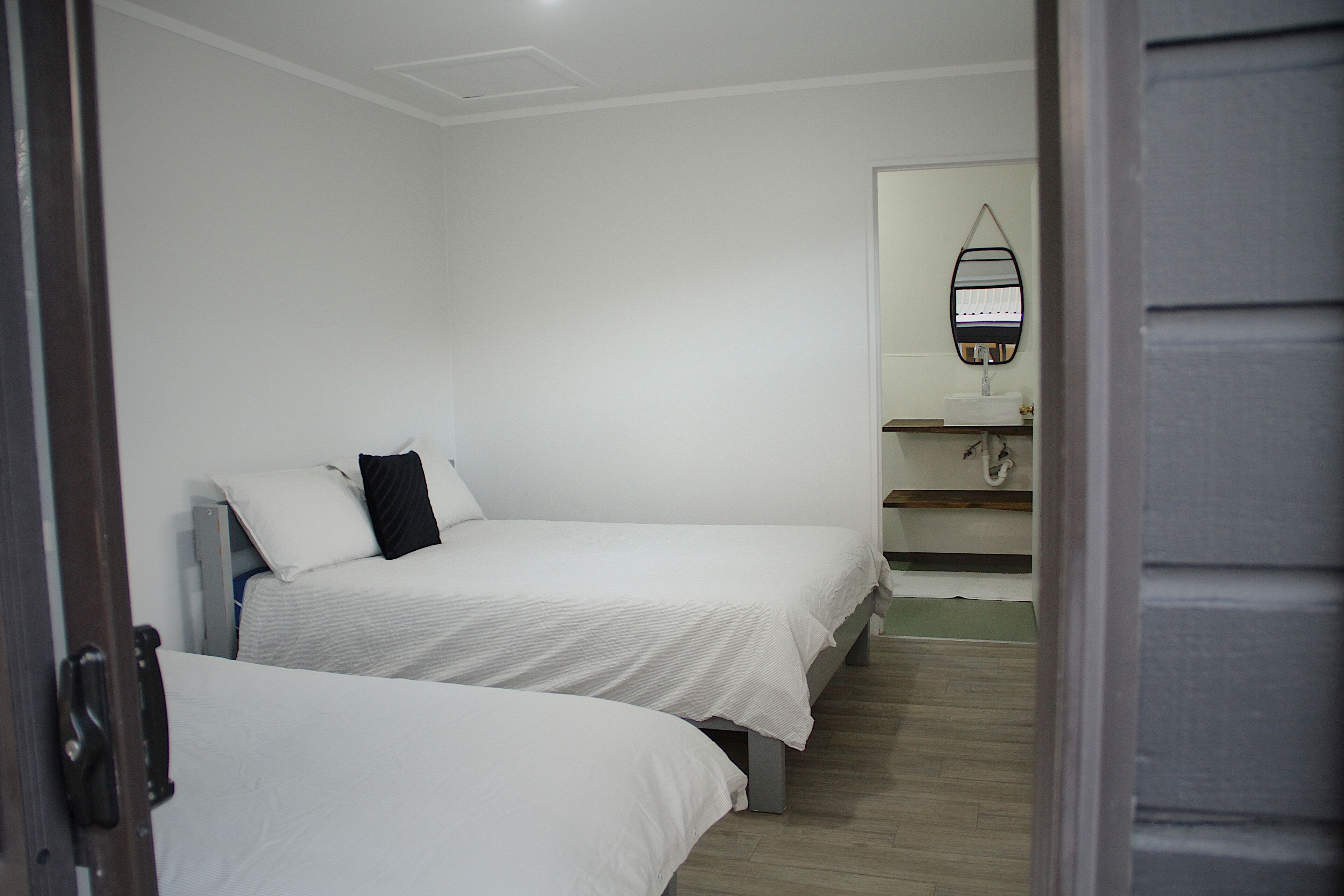 A bedroom with two single beds, a small bathroom with a mirror and sink, and a wooden floor.