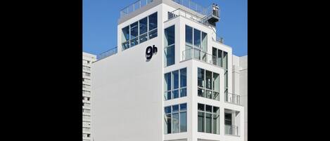 A modern building with a prominent '9h' sign, multiple balconies, and a rooftop terrace.