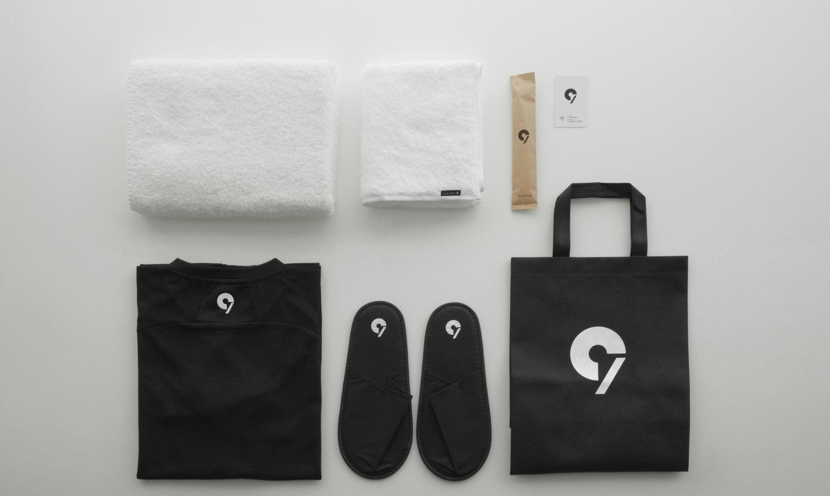 A black tote bag, a pair of black slippers, a black t-shirt, two white towels, a brown paper bag, and a card with a logo.