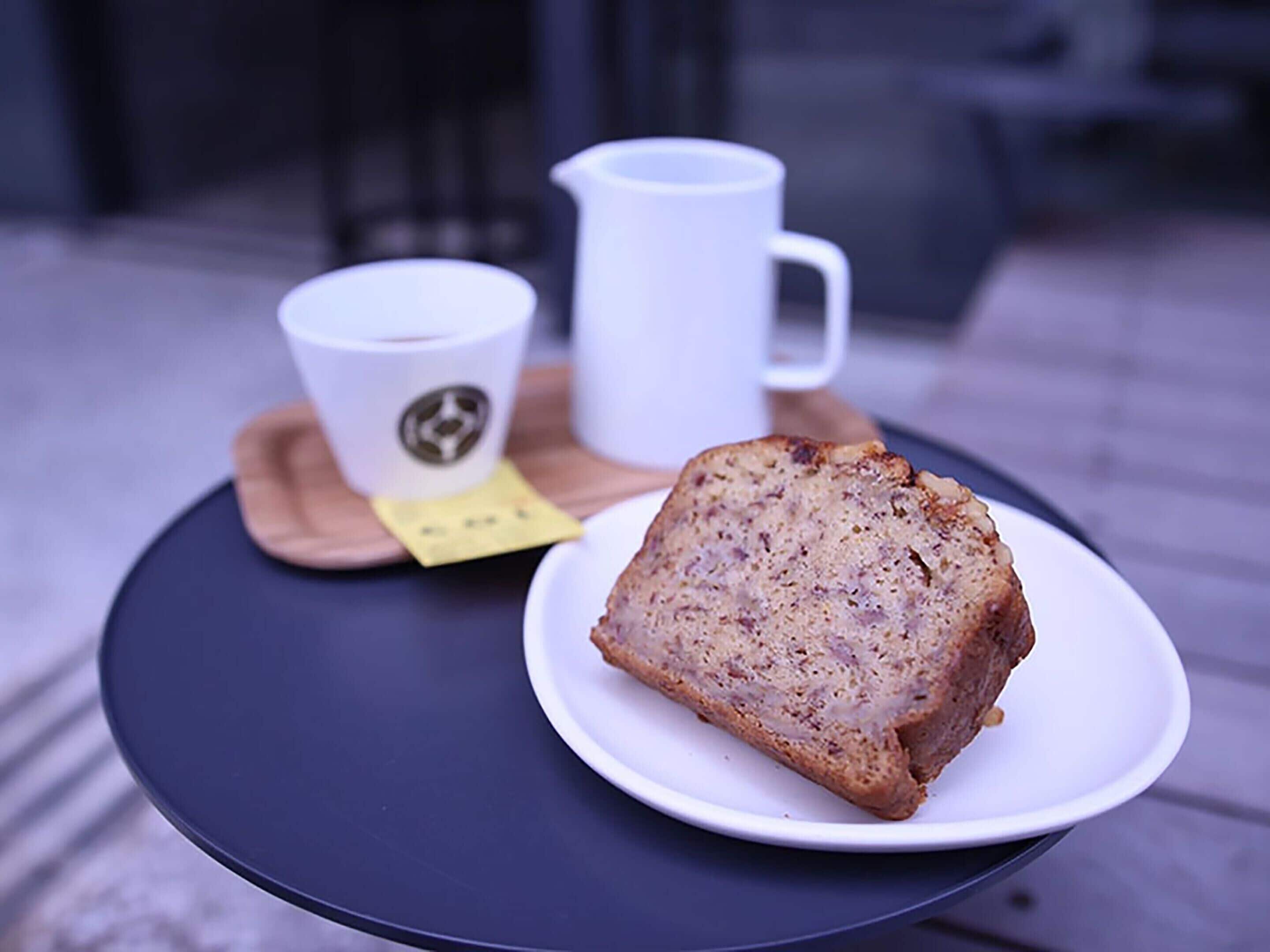 A slice of bread on a white plate on a round table, a white mug, and a coaster with a logo.