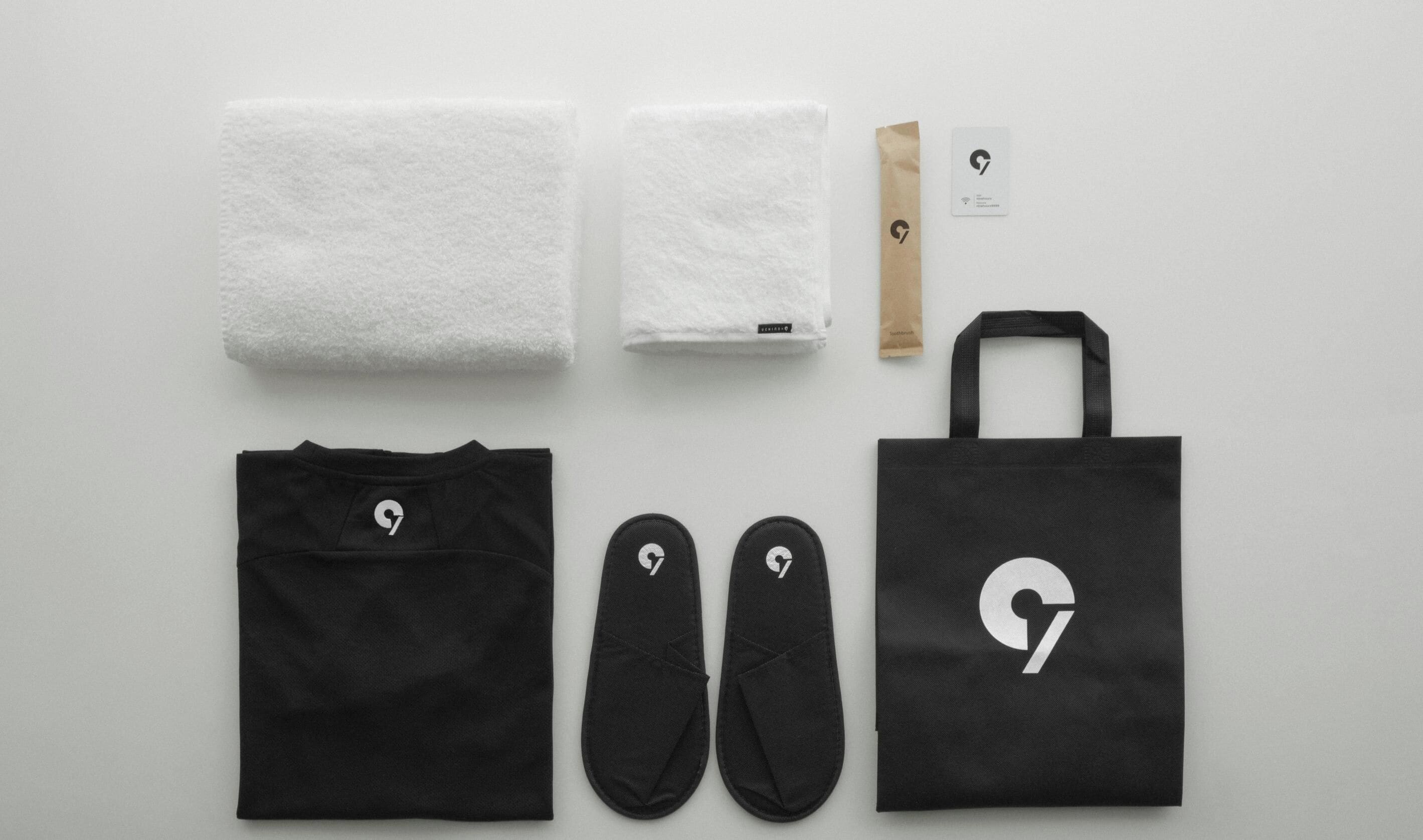 A black tote bag, a pair of black slippers, a black t-shirt, two white towels, a brown paper bag, and a card with a logo.
