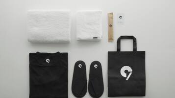 A black tote bag, a pair of black slippers, a black t-shirt, two white towels, a brown paper bag, and a card with a logo.