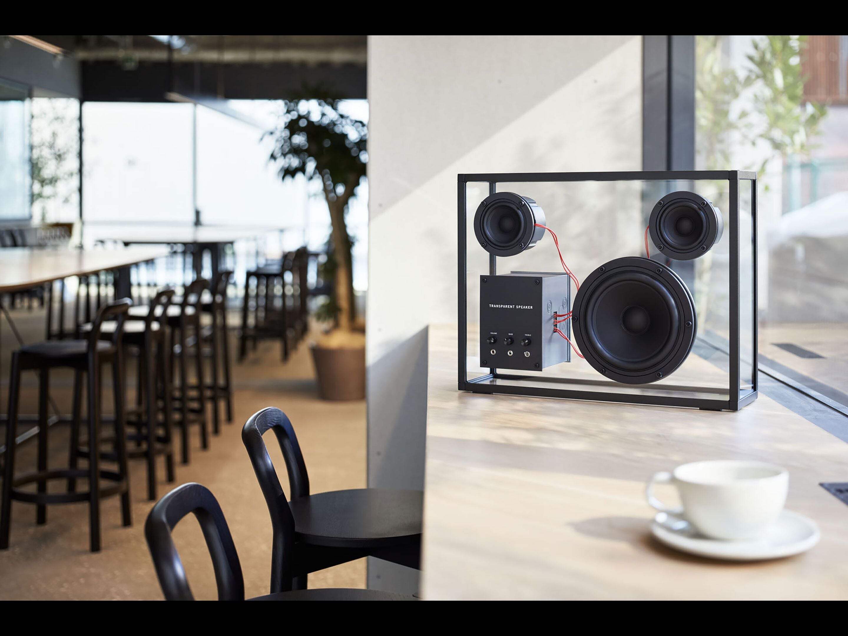 Ein modernes Café mit minimalistischer Einrichtung, das eine Glasvitrine mit einem transparenten Lautsprechersystem und einer weißen Tasse auf Untertasse zeigt.