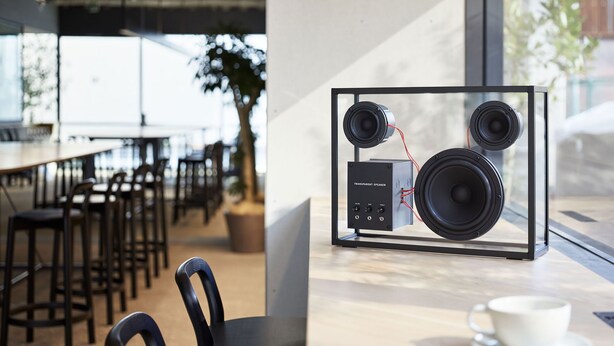 Un café moderno con una decoración minimalista, que incluye un expositor de vidrio con un sistema de altavoces transparente y una taza blanca sobre un platito.