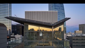 Un edificio moderno con fachada de vidrio, una terraza en la azotea con suelo de madera y vistas del horizonte urbano al atardecer.