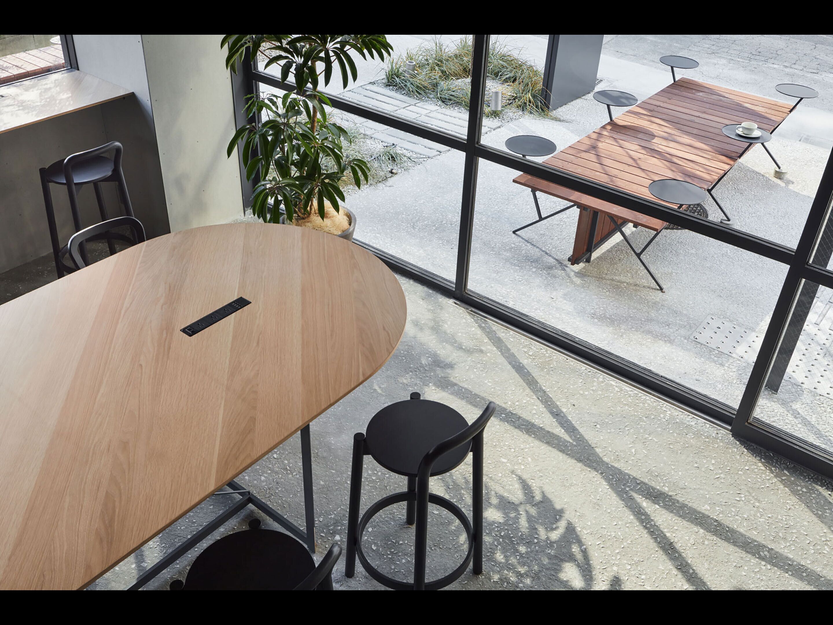 Un salón de conferencias con una mesa de madera y sillas negras, ventanas amplias con vistas a la calle y un área de estar al aire libre.