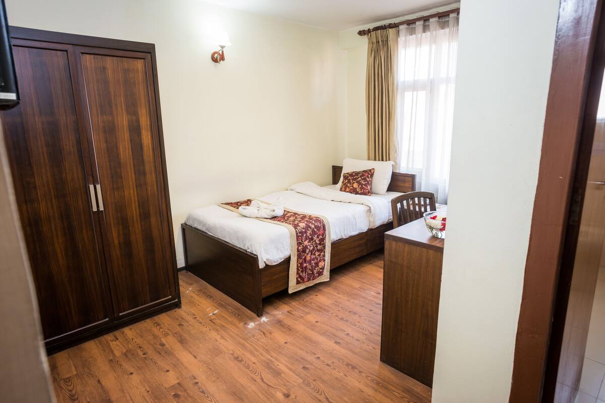 A bedroom with a wooden wardrobe, a bed with a floral bedspread, and a bedside table with a vase.