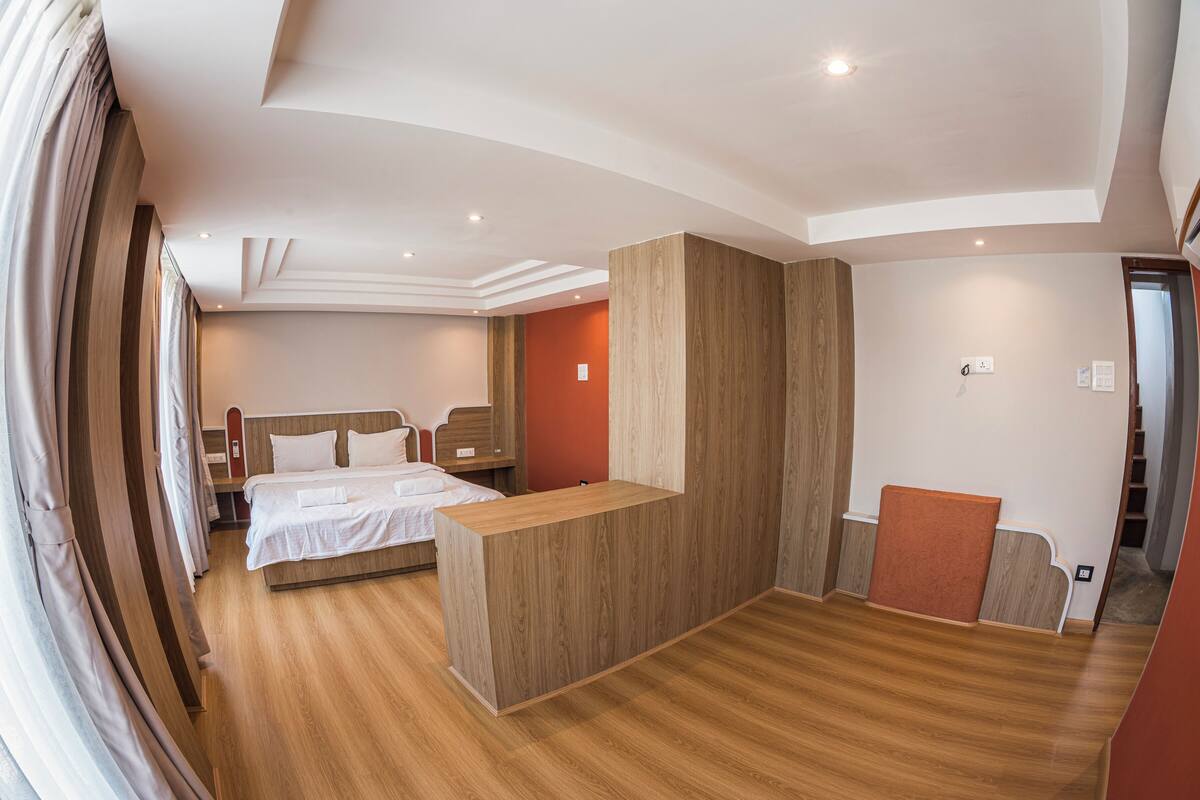 A hotel room with a wooden floor, a bed with white linens, a wooden reception desk, and a red accent wall.