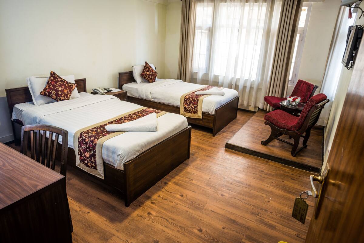 A hotel room with two beds, wooden flooring, a small table with a chair, and a door with a handle.