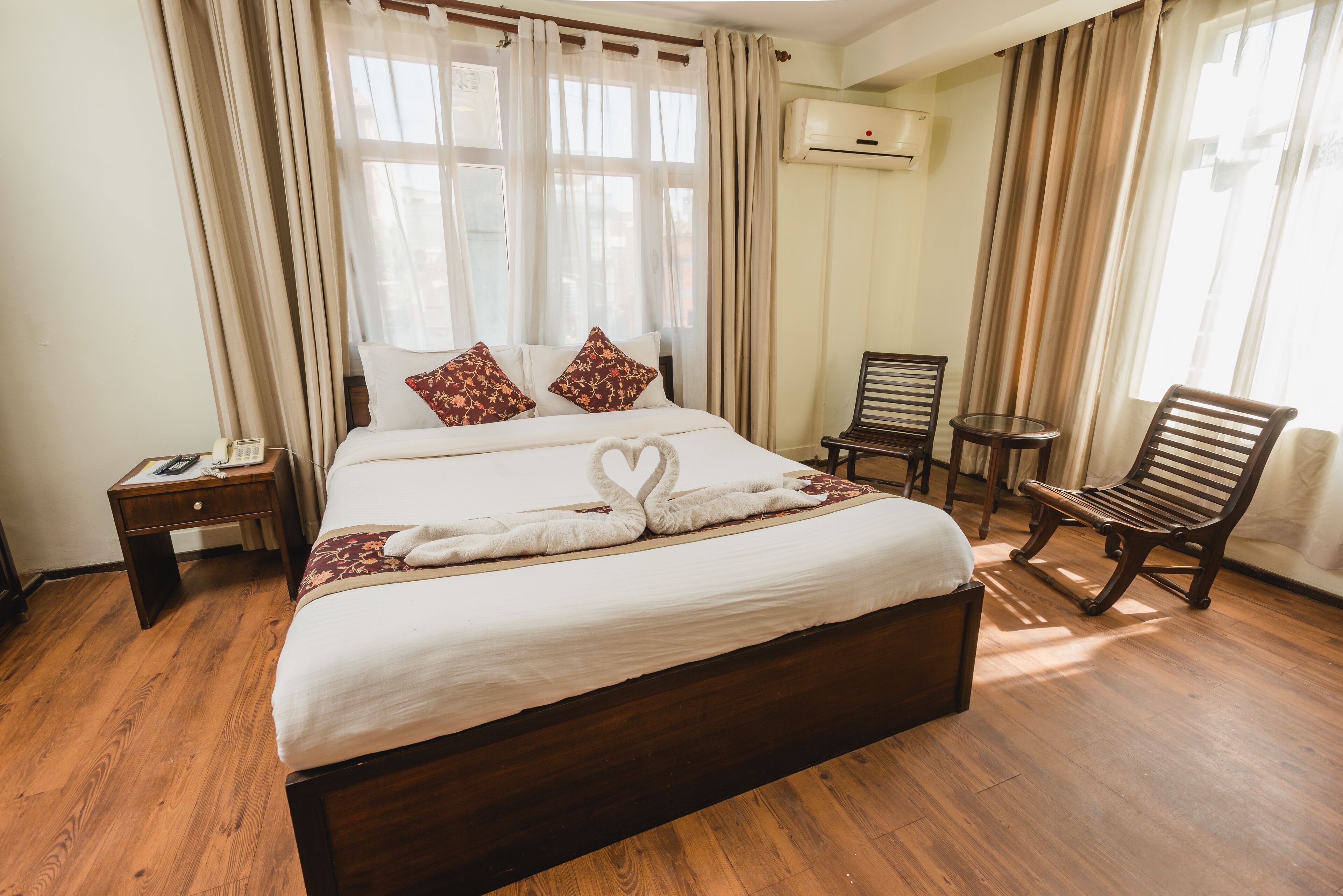 A hotel room with a large bed, two pillows, a bedside table with a phone, a small round table with chairs, and an air conditioning unit on the wall.