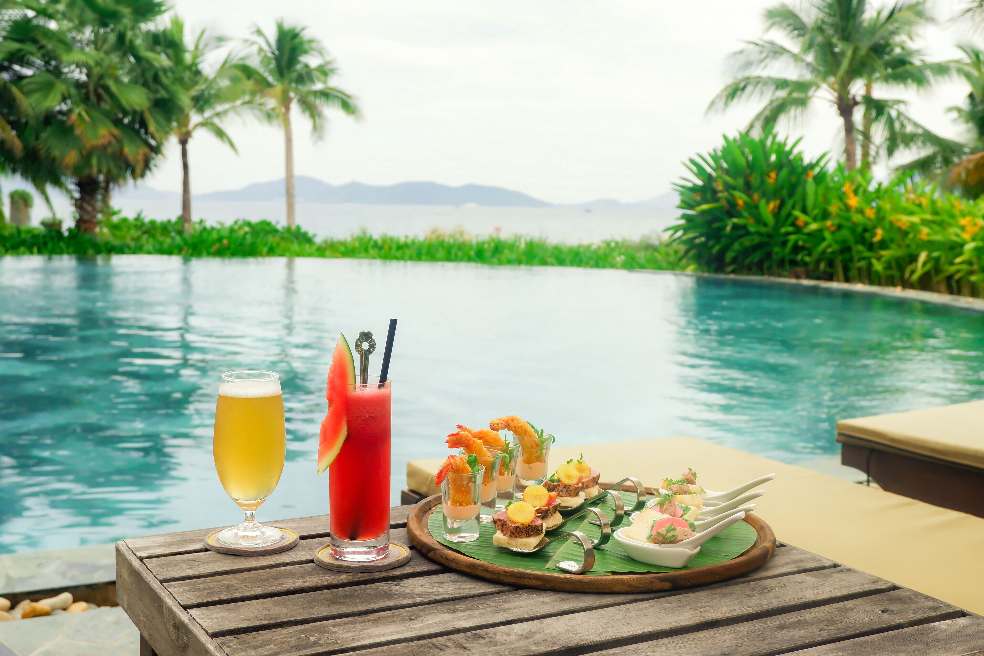 A wooden table with a glass of beer, a cocktail, and a tray of appetizers by a pool with palm trees and mountains in the background.