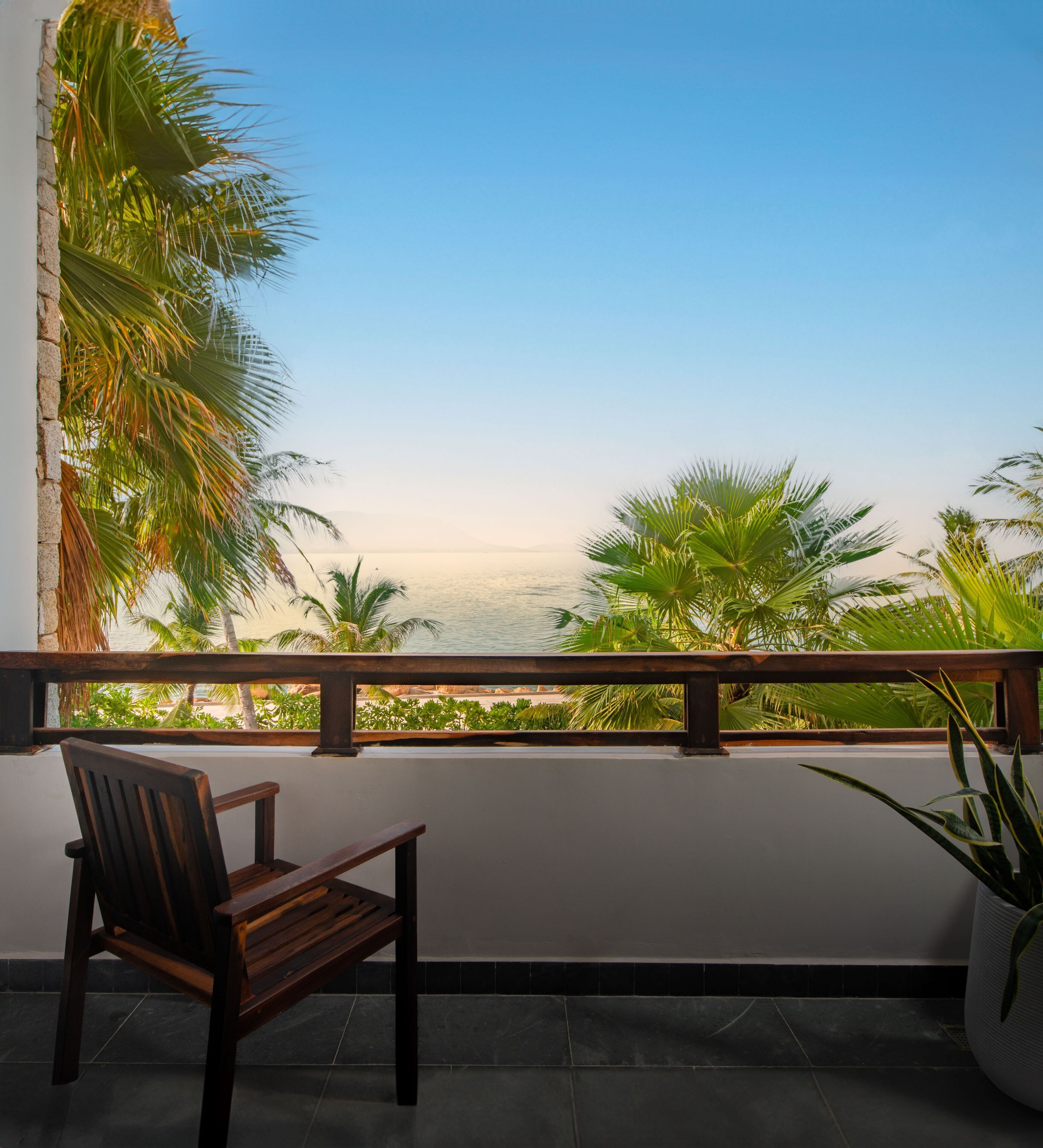 Un balcon avec une chaise en bois, un pot de fleurs et une vue sur les palmiers et la mer.