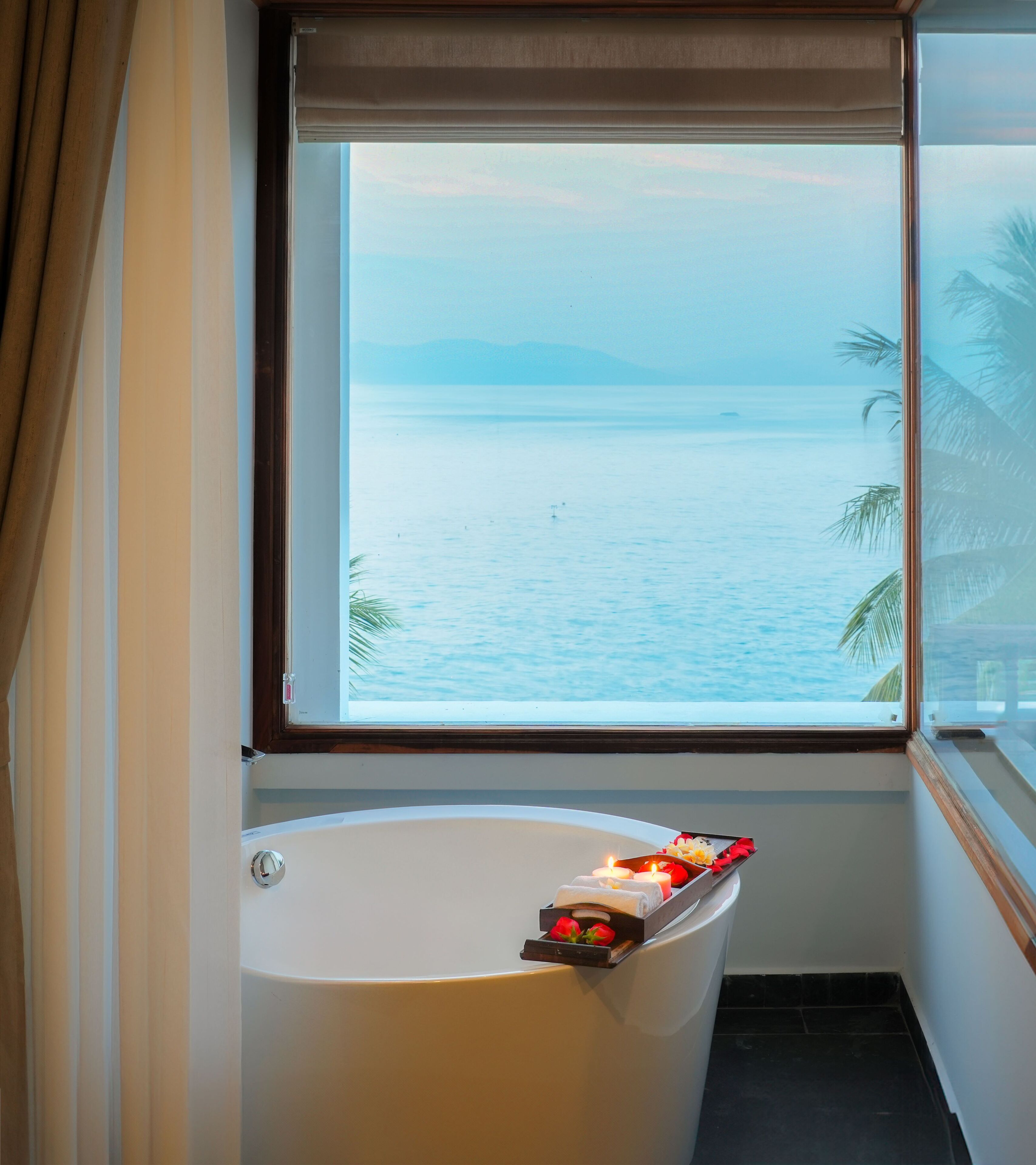 Una bañera con una bandeja de artículos de aseo y una ventana que ofrece vistas a un mar tranquilo y montañas distantes.