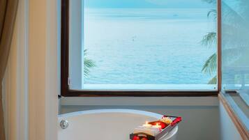 Une baignoire avec un plateau contenant des articles de toilette et une fenêtre donnant sur une mer calme et des montagnes lointaines.